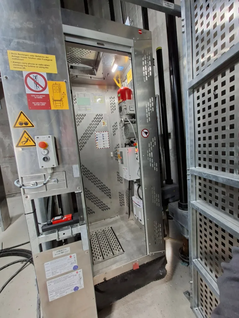 Industrial stainless-steel machine enclosure elevator with an open access door, control panel, emergency stop button, warning signs, and surrounding metal safety guards