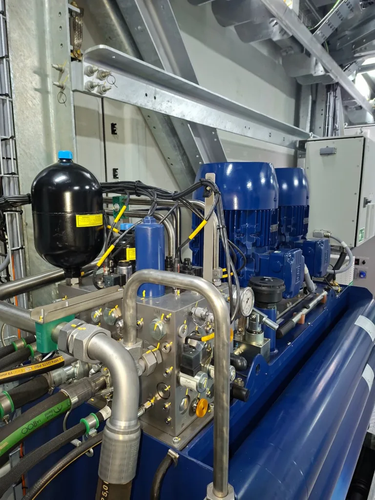 Industrial hydraulic system with blue electric motors, pressure accumulator, metal piping, hoses, valves, and control components.